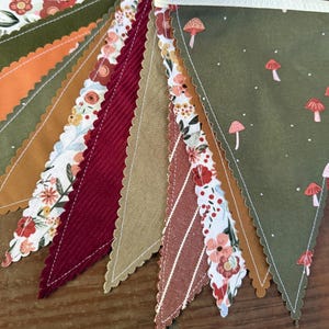 May include: A colorful bunting banner with various patterned pennants. The designs include floral prints, corduroy, and mushroom motifs in shades of green, orange, red, and tan. The edges are scalloped and sewn with white thread.
