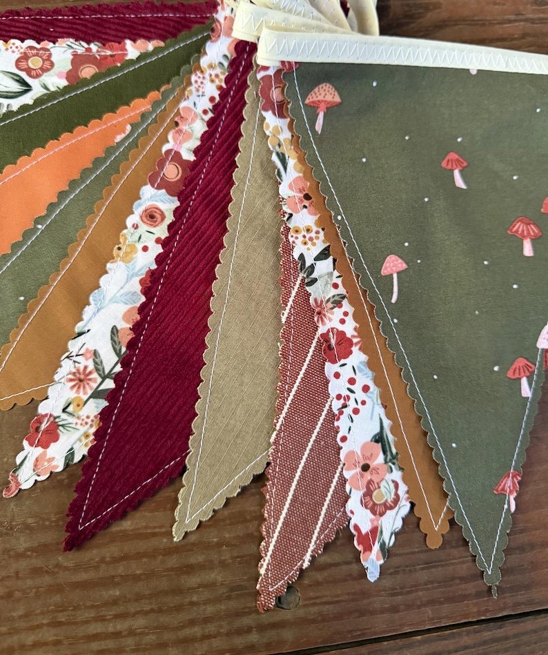 May include: A colorful bunting banner with various patterned pennants. The designs include floral prints, corduroy, and mushroom motifs in shades of green, orange, red, and tan. The edges are scalloped and sewn with white thread.