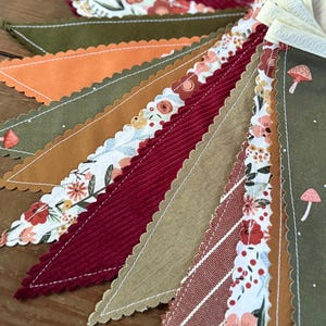 May include: A close-up of a colorful fabric banner with a variety of patterns and textures. The banner features triangles in shades of red, orange, green, and tan, with floral and mushroom designs. The edges are scalloped and stitched with white thread.