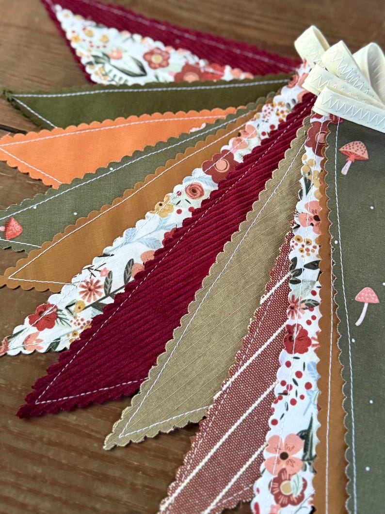 May include: A close-up of a colorful fabric banner with a variety of patterns and textures. The banner features triangles in shades of red, orange, green, and tan, with floral and mushroom designs. The edges are scalloped and stitched with white thread.