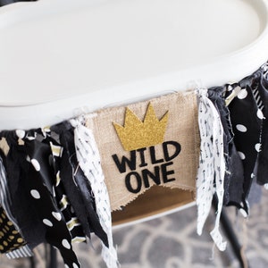 May include: A white high chair with a black and white patterned seat and a hessian banner that says "Wild One" with a gold glitter crown on top. The banner is decorated with black and white fabric scraps.