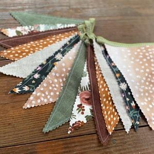 May include: A fabric banner with a scalloped edge, featuring a variety of patterns in shades of brown, green, and white. The banner is made of different fabrics, including linen and cotton, and features floral, polka dot, and geometric designs.