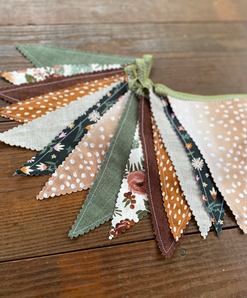 May include: A fabric banner with a scalloped edge, featuring a variety of patterns in shades of brown, green, and white. The banner is made of different fabrics, including linen and cotton, and features floral, polka dot, and geometric designs.