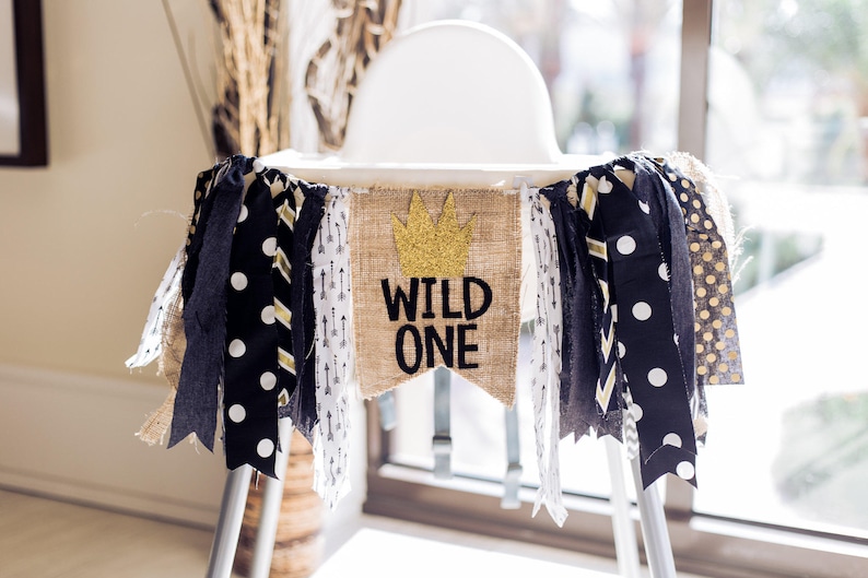 May include: A burlap banner with the words "Wild One" and a gold crown on a white high chair decorated with black and white polka dot ribbon, black and white chevron ribbon, and gold and white ribbon.