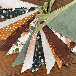 May include: A bunting banner with 12 triangular flags in various shades of brown, green, and white. The flags are decorated with different patterns, including polka dots, floral designs, and solid colors. The bunting is perfect for adding a touch of rustic charm to any party or event.