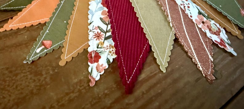 May include: A close-up of a decorative banner with various fabric pennants. The pennants feature a variety of colors and patterns, including orange, green, floral, and red. Each pennant has a scalloped edge and is arranged in a row.