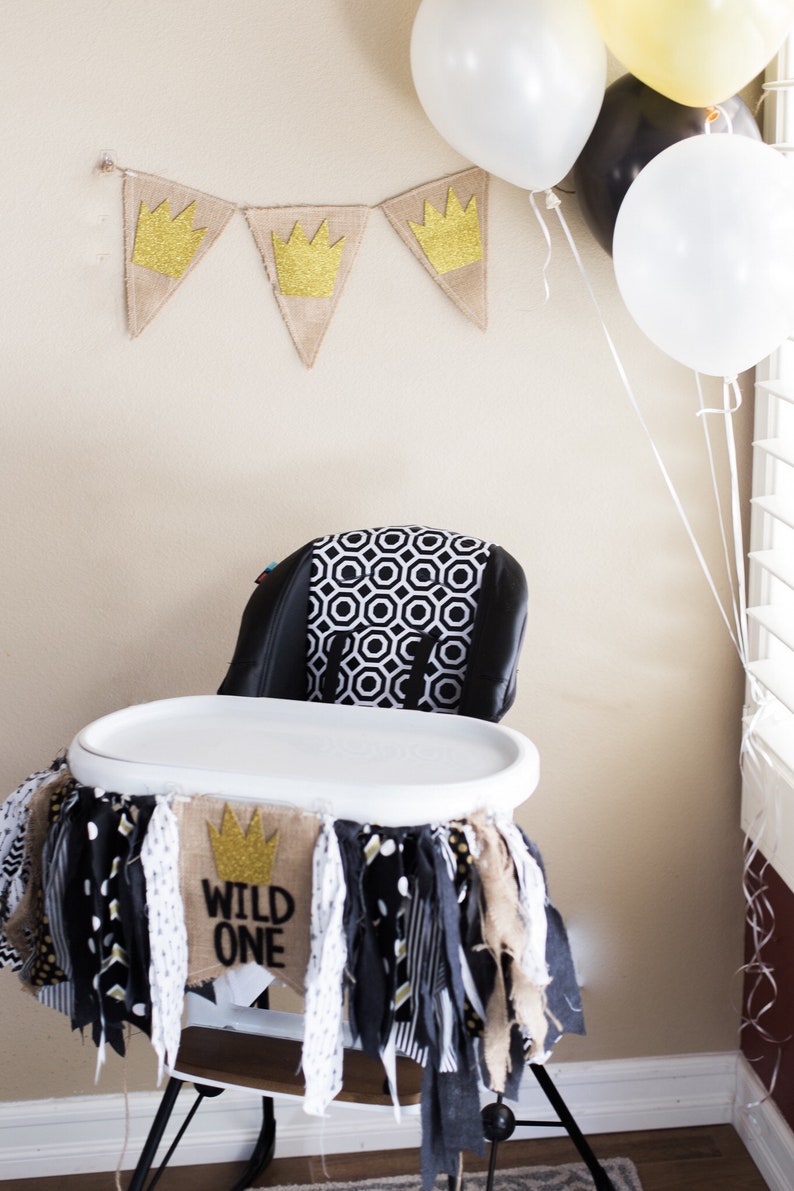 May include: A white high chair with a black and white patterned seat cushion. The chair is decorated with a hessian banner that says "Wild One" and has a gold crown. The banner is surrounded by black, white and gold fabric scraps.