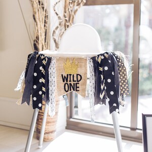 May include: A white high chair with a burlap banner that says "Wild One" in black letters with a gold crown above the text. The banner is decorated with black and white polka dot fabric and gold and white chevron fabric.
