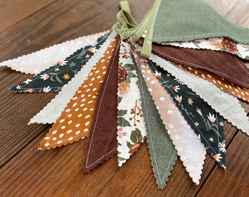 May include: A bunting banner with a variety of fabric flags in different patterns and colors, including green, brown, white, and orange. The flags have a scalloped edge and are sewn together with white thread.
