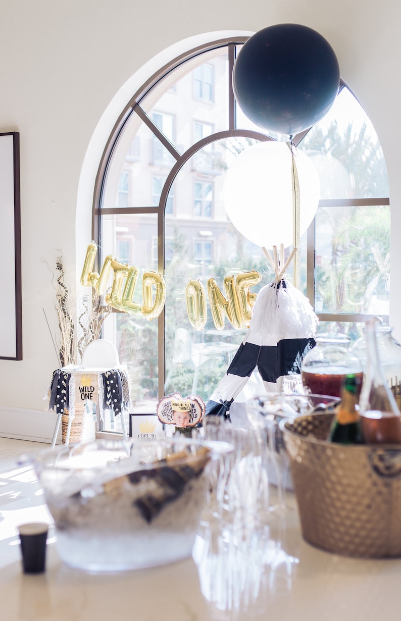 May include: A white and black teepee tent with a white feather trim sits in front of a large window with gold foil balloons spelling "WILD ONE".  There are black and white balloons and a table with a glass bowl filled with ice and a gold bucket with champagne bottles.