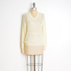 Puede incluir: Un suéter blanco de crochet con cuello en V y un patrón geométrico. El suéter está en un maniquí.