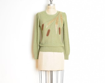 Könnte beinhalten: Ein grüner Pullover mit einem braunen und goldenen Stickmuster aus Weizenähren. Der Pullover befindet sich auf einer weißen Schaufensterpuppe.