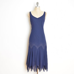 May include: A navy blue dress with a V-neck and a flowing, asymmetrical hemline. The dress features a beaded embellishment at the waist and a layered skirt. The dress is displayed on a vintage dress form.