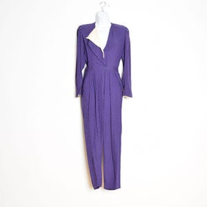 Puede incluir: Mono morado vintage con un sutil estampado de lunares. La prenda de manga larga presenta un cuello en V con solapa y puños color crema. El mono está colgado de una percha blanca contra una pared blanca.