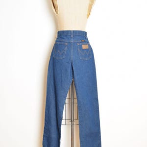 May include: A pair of blue denim jeans displayed on a mannequin. The jeans have a high waist, classic five-pocket design, and a brown leather patch on the back. The Wrangler logo is visible on the patch and back pockets.
