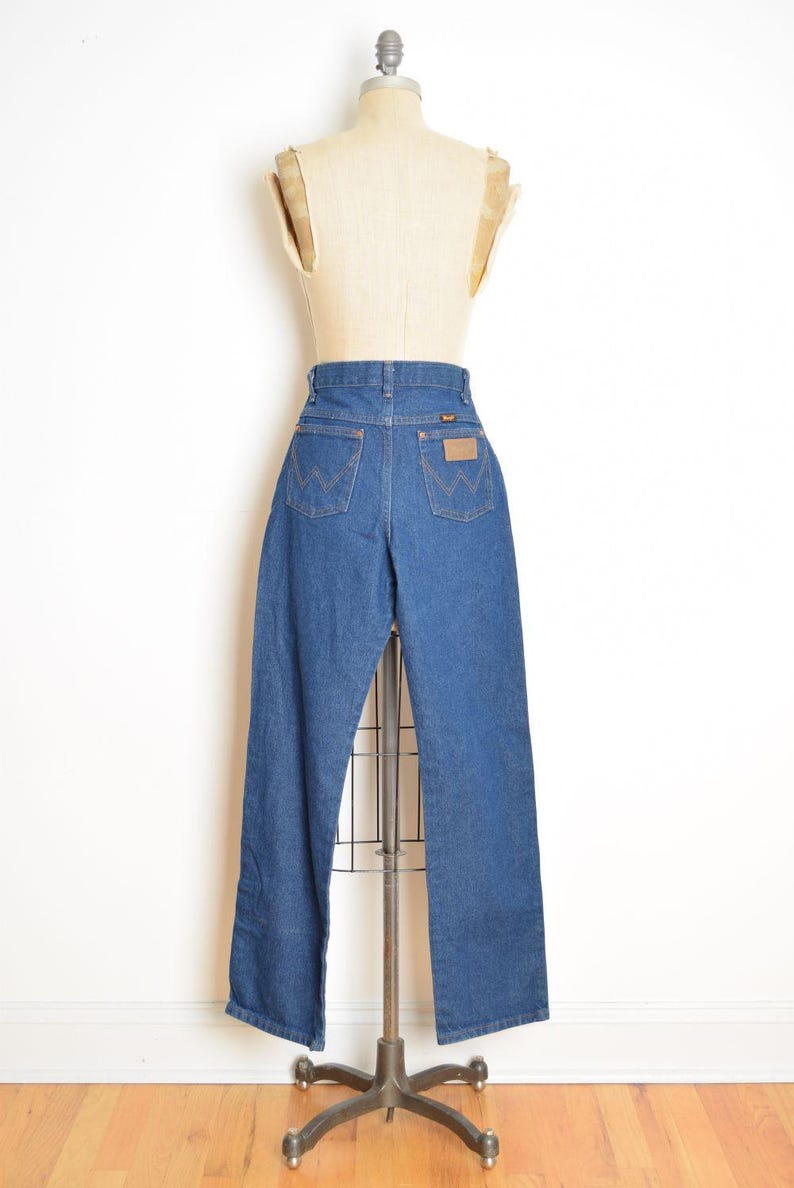 May include: A pair of blue denim jeans displayed on a mannequin. The jeans have a high waist, classic five-pocket design, and a brown leather patch on the back. The Wrangler logo is visible on the patch and back pockets.