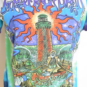 May include: White t-shirt with a colorful graphic featuring a lighthouse, skeleton in a boat, and the words "Grateful Dead" in a stylized font. The design includes a sun, waves, and a tie-dye effect on the sleeves.