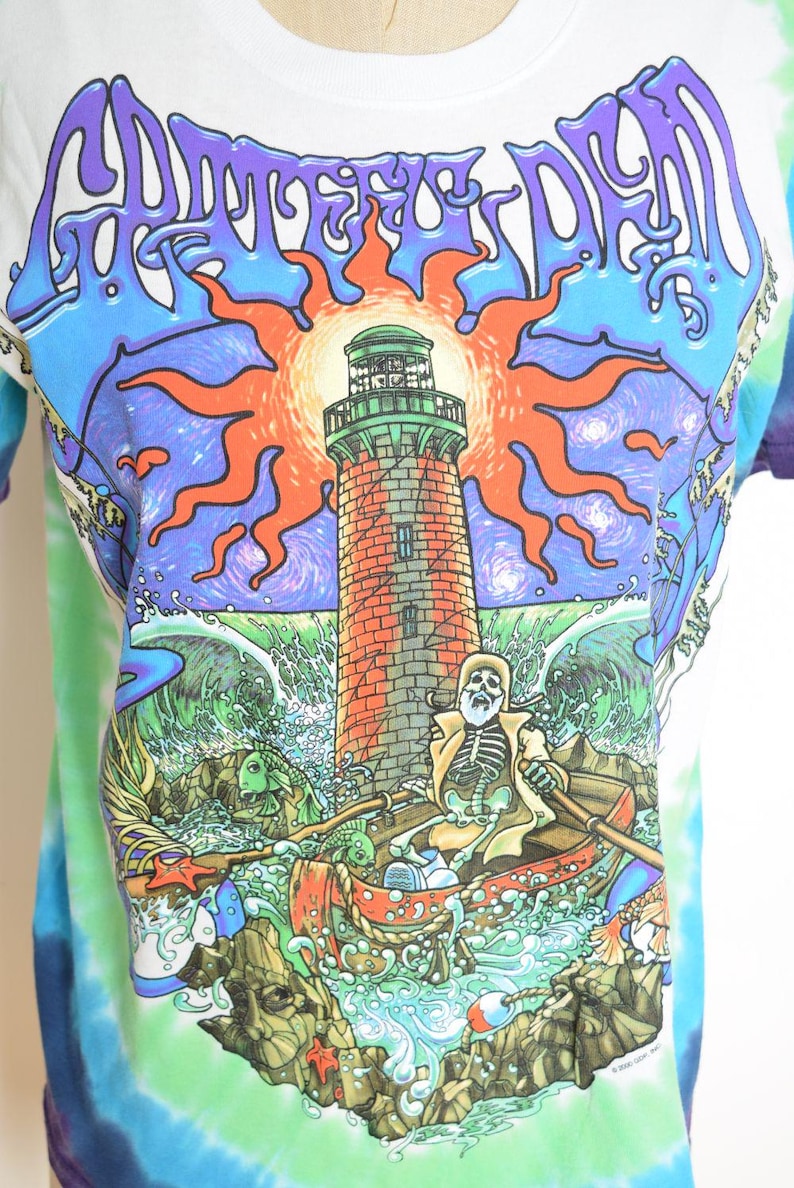 May include: White t-shirt with a colorful graphic featuring a lighthouse, skeleton in a boat, and the words "Grateful Dead" in a stylized font. The design includes a sun, waves, and a tie-dye effect on the sleeves.