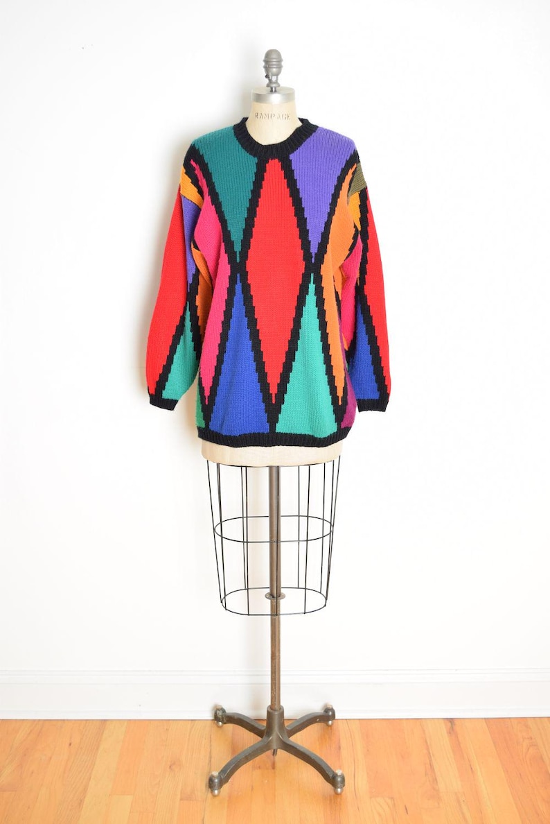 May include: A vintage sweater with a geometric pattern in red, pink, green, blue, purple, and orange. The sweater has a black crew neck and black trim. The sweater is displayed on a vintage dress form.