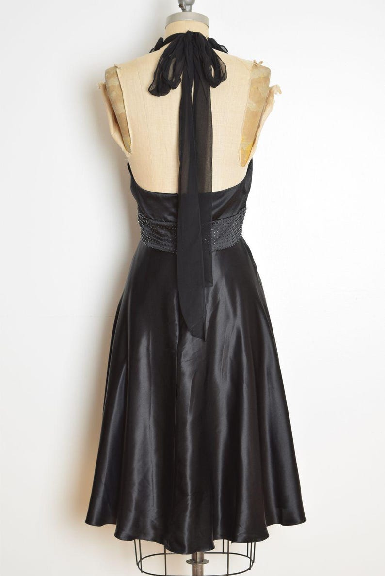 May include: A black satin halter dress with a beaded waistline and a flowing skirt. The dress features a long, sheer tie that drapes down the back. The dress is displayed on a mannequin.