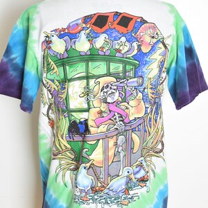 May include: White tie-dye t-shirt with a colorful graphic. The graphic features a skeleton with a telescope, surrounded by cartoon birds and a vibrant, surreal landscape. The shirt has blue, green, and purple tie-dye on the sleeves.