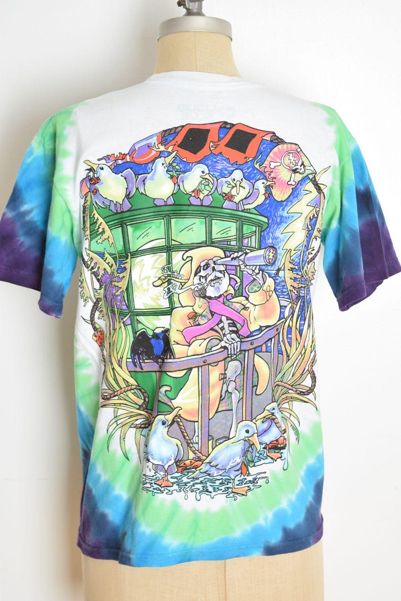 May include: White tie-dye t-shirt with a colorful graphic. The graphic features a skeleton with a telescope, surrounded by cartoon birds and a vibrant, surreal landscape. The shirt has blue, green, and purple tie-dye on the sleeves.
