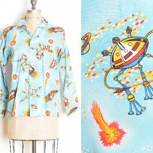 May include: Light blue button-down shirt with a space-themed print. The shirt features a repeating pattern of rockets, planets, and other space-related imagery in shades of yellow, orange, and white. The shirt is displayed on a mannequin.