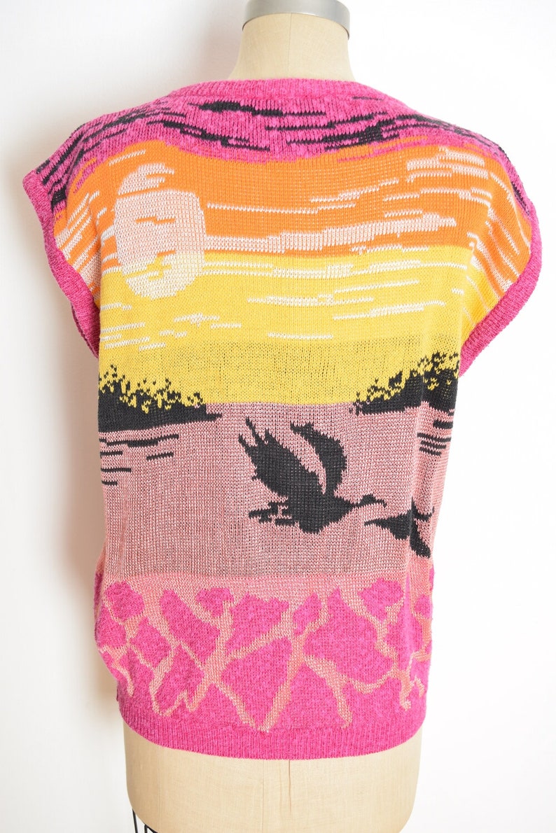 May include: A pink sleeveless sweater with a sunset and birds design. The sweater has a black, orange, yellow, and pink color scheme.