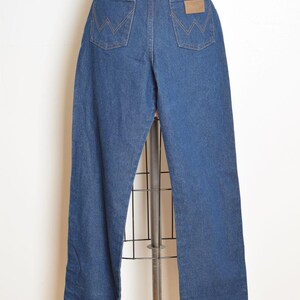 May include: Blue denim Wrangler jeans displayed on a mannequin. The jeans have a high waist, straight leg, and classic five-pocket design. The back pockets feature the signature Wrangler "W" stitching, and a brown leather patch is visible.