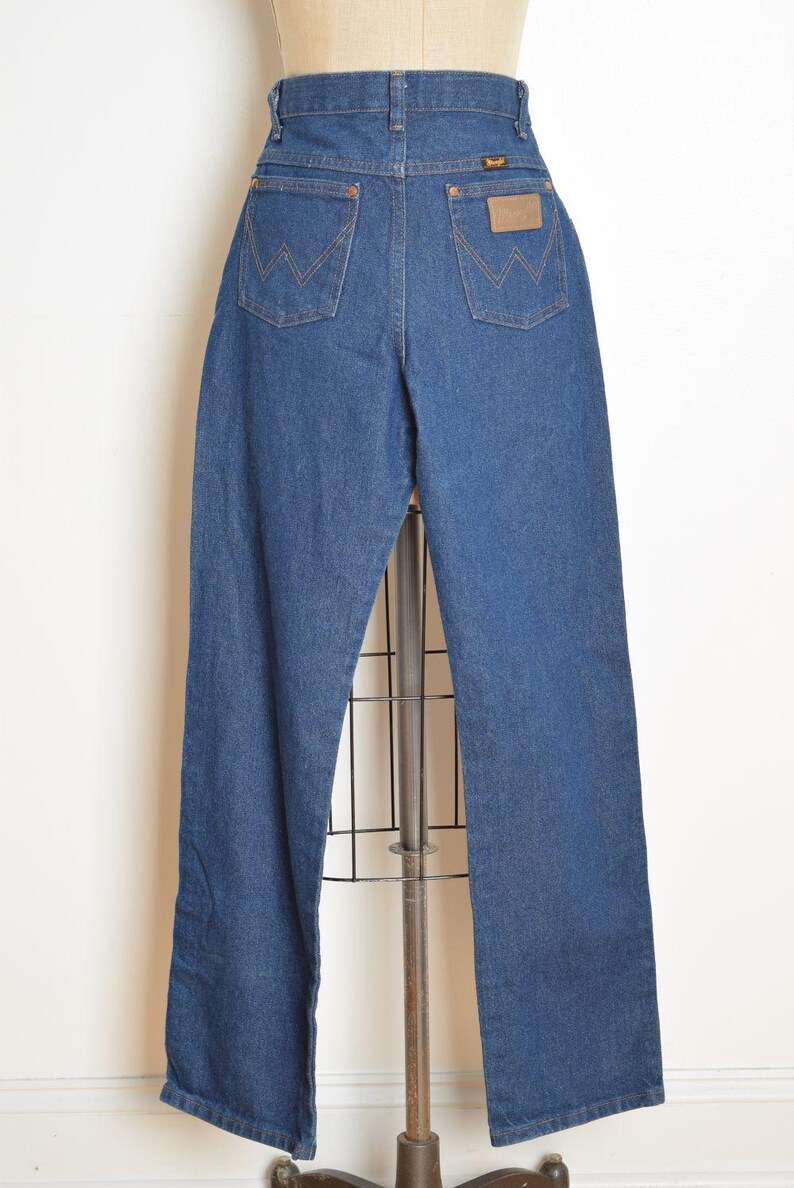 May include: Blue denim Wrangler jeans displayed on a mannequin. The jeans have a high waist, straight leg, and classic five-pocket design. The back pockets feature the signature Wrangler "W" stitching, and a brown leather patch is visible.