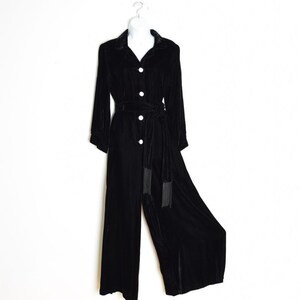 Puede incluir: Un mono de terciopelo negro con mangas largas, cuello camisero y cierre frontal de botones. La prenda incluye un cinturón de lazo a juego en la cintura con detalles de flecos en los extremos y pantalones de pierna ancha hasta el suelo.