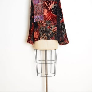 May include: A patchwork velvet top with long sleeves, displayed on a dress form. The top features a mix of floral and geometric patterns in shades of red, black, purple, and peach. The garment has a relaxed fit.