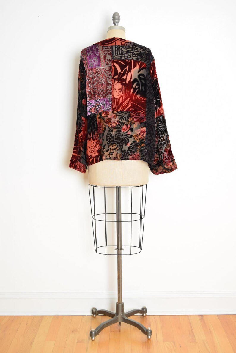 May include: A patchwork velvet top with long sleeves, displayed on a dress form. The top features a mix of floral and geometric patterns in shades of red, black, purple, and peach. The garment has a relaxed fit.
