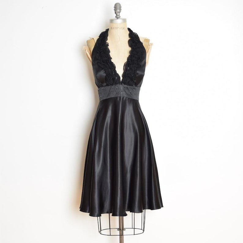 May include: A black satin halter dress with a flared skirt. The bodice features a ruffled neckline and a beaded waist. The dress is displayed on a mannequin against a white background. The dress is knee-length.