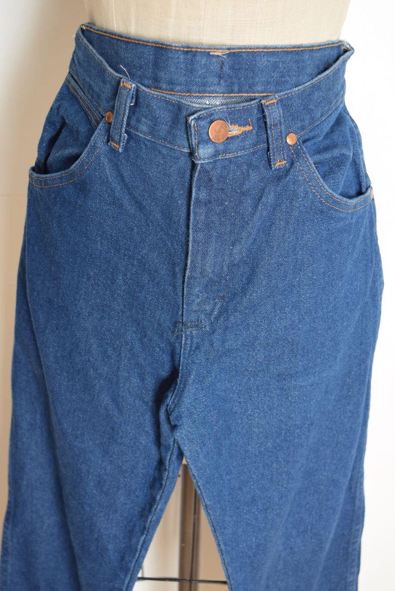 May include: Close-up of blue denim jeans. The jeans have a high waist, copper-colored rivets, and a button closure. The pockets are visible, and the stitching is in a contrasting color. The denim appears to be a medium wash.