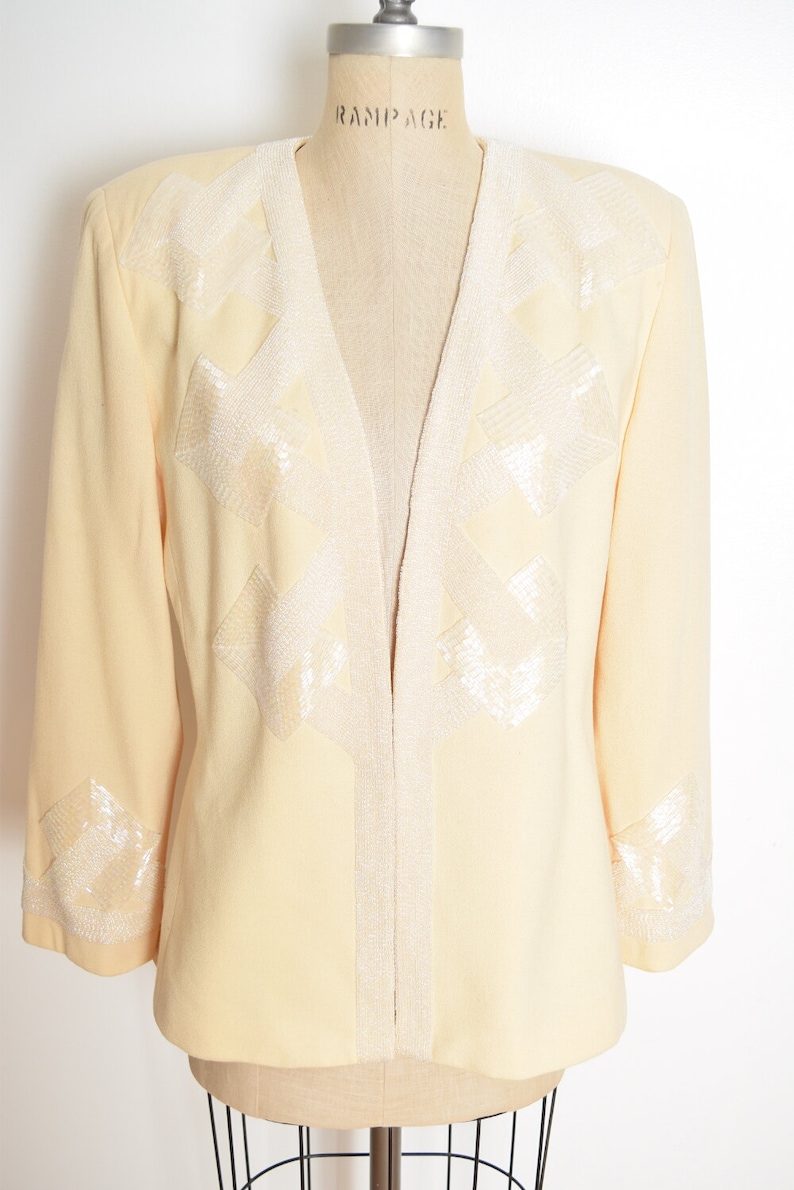 May include: A pale yellow blazer with a V-neck and long sleeves. The blazer is decorated with white beaded geometric designs on the front and cuffs. The label "Rampage" is visible on the mannequin's chest.