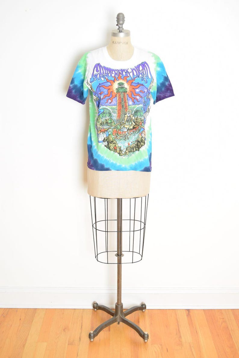 May include: White tie-dye t-shirt with a colorful graphic featuring a lighthouse, sun, and text. The shirt is displayed on a vintage dress form. The sleeves are blue and purple.