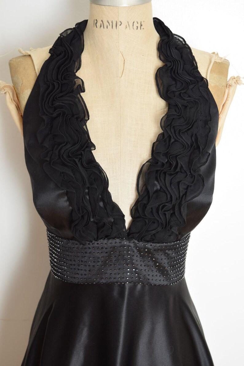 May include: Black halter dress with a ruffled neckline and a beaded waist. The dress is made of a shiny, satin-like material. The dress is displayed on a mannequin.