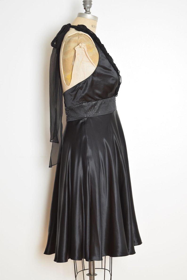 May include: A black satin dress with a halter neckline and a beaded belt. The dress has a flowing skirt and a sheer, draped accent over one shoulder. The dress is displayed on a mannequin.