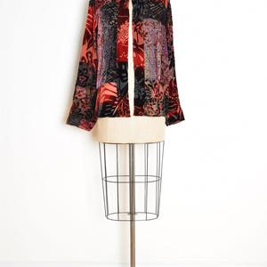May include: A long-sleeved, open-front jacket with a patchwork design. The jacket features a mix of red, black, and patterned fabrics. It is displayed on a vintage dress form with a wire cage base.