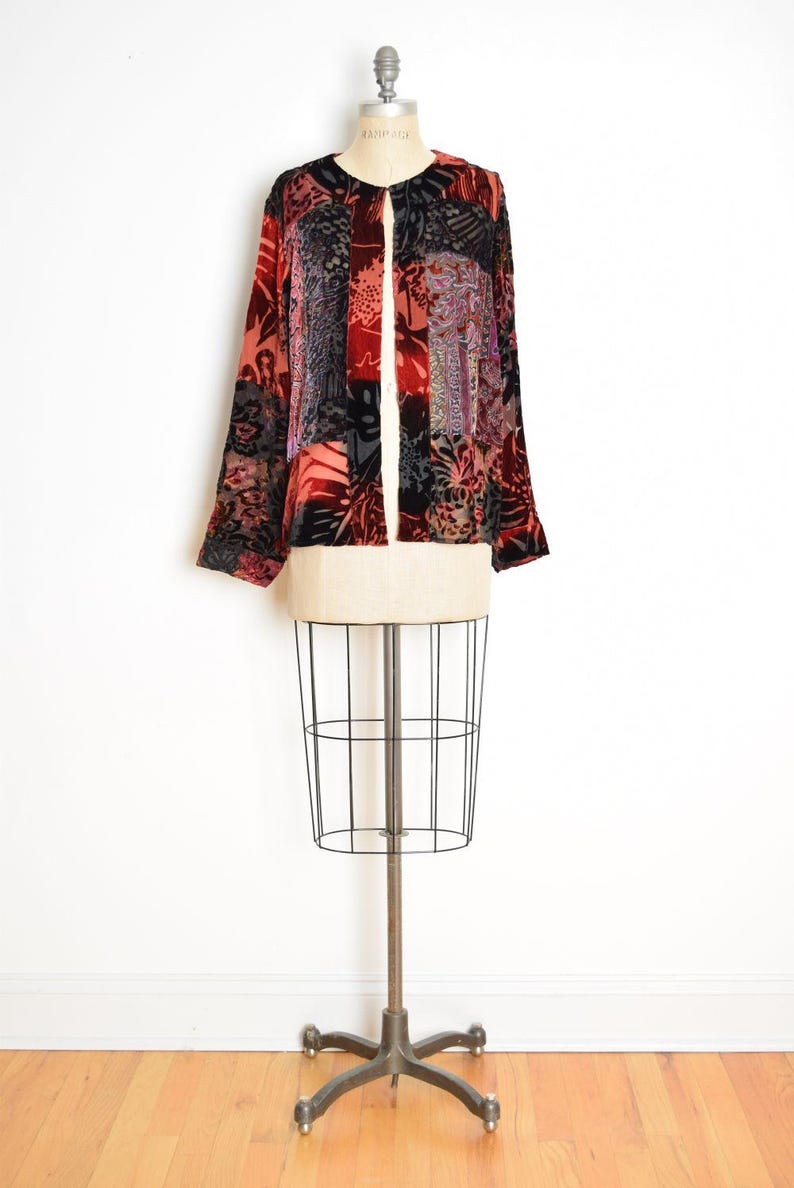 May include: A long-sleeved, open-front jacket with a patchwork design. The jacket features a mix of red, black, and patterned fabrics. It is displayed on a vintage dress form with a wire cage base.