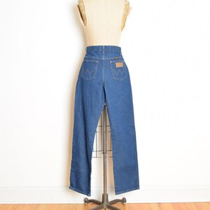 May include: Blue denim jeans displayed on a mannequin. The jeans have a high waist, a straight leg, and classic five-pocket styling. A brown leather patch is visible on the back. The jeans are displayed on a metal stand.