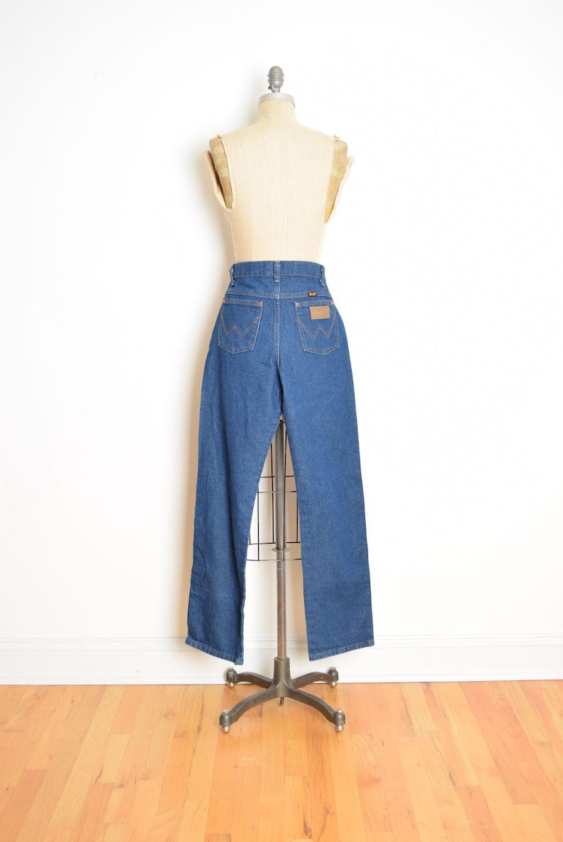 May include: Blue denim jeans displayed on a mannequin. The jeans have a high waist, a straight leg, and classic five-pocket styling. A brown leather patch is visible on the back. The jeans are displayed on a metal stand.
