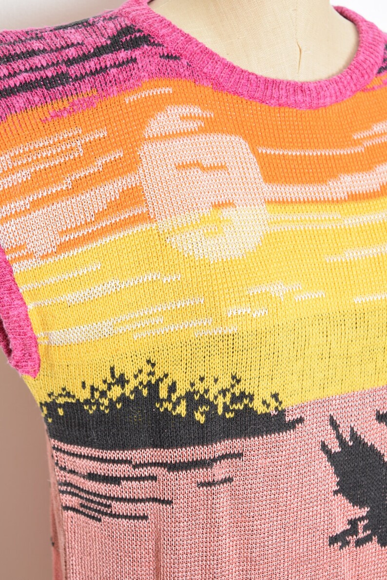 May include: A sleeveless knitted sweater with a colorful sunset design. The sweater is pink, orange, yellow, and black. The design features a sun and a palm tree.