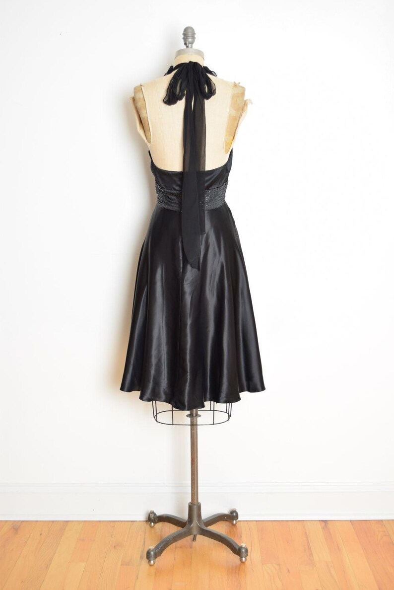 May include: A black satin halter dress displayed on a vintage dress form. The dress features a fitted bodice, a beaded waist detail, and a flowing A-line skirt. A long black tie extends from the halter neckline.