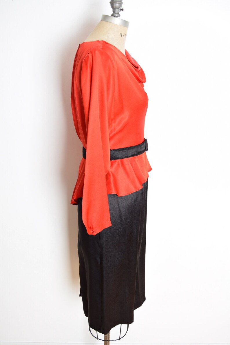 May include: A red and black dress with a cowl neck and a black belt. The dress has long sleeves and a skirt that falls to the knees.