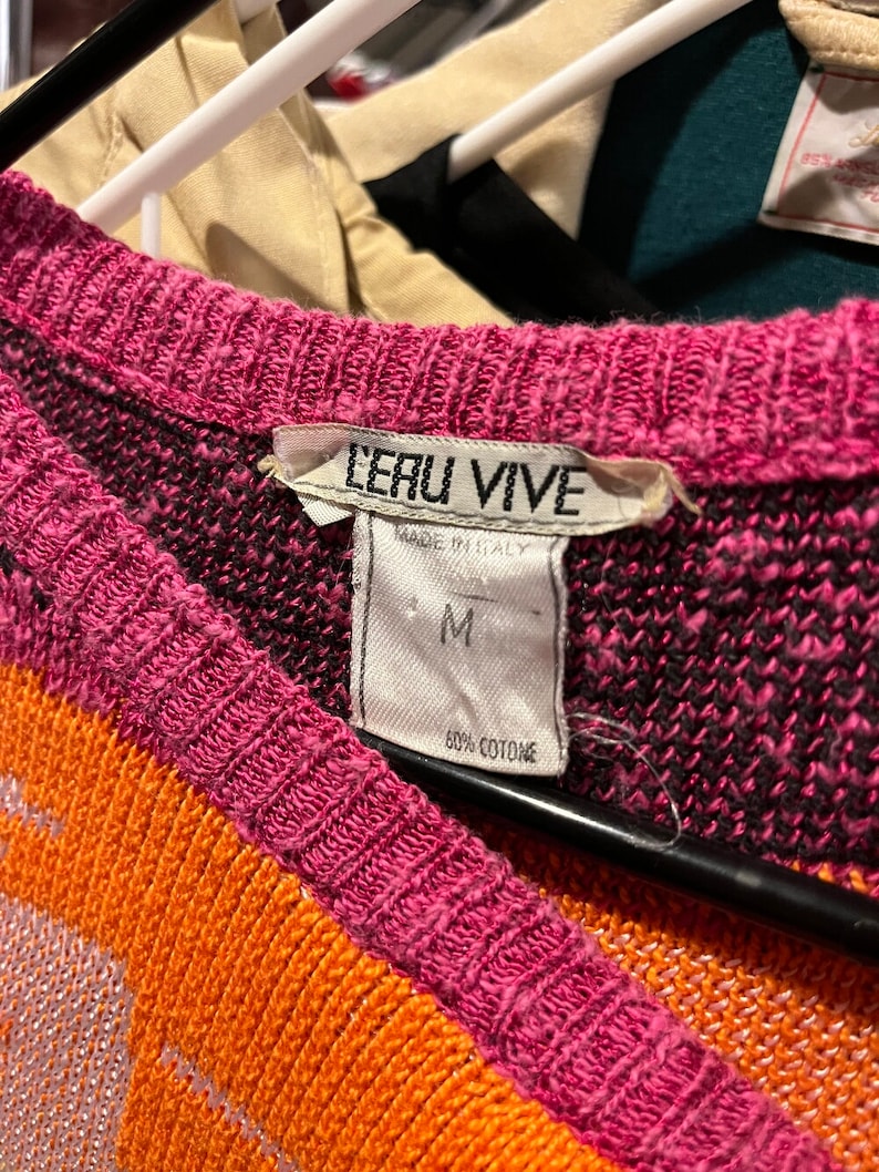 May include: A pink, orange, and black patterned sweater with a white tag that reads "LERU VIVE" and "MADE IN ITALY". The tag also indicates the size "M" and "60% COTONE".