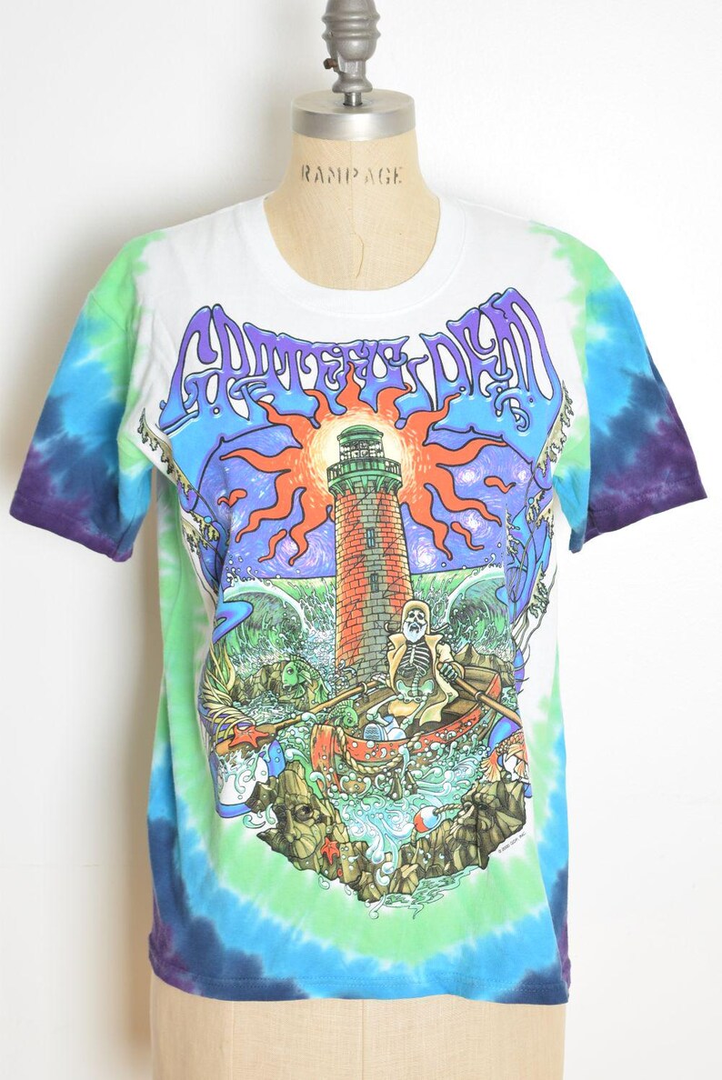 May include: White tie-dye t-shirt with a colorful graphic featuring a lighthouse, skeleton, and boat. The shirt has a crew neck and short sleeves. The graphic includes the text "Grateful Dead" in purple.