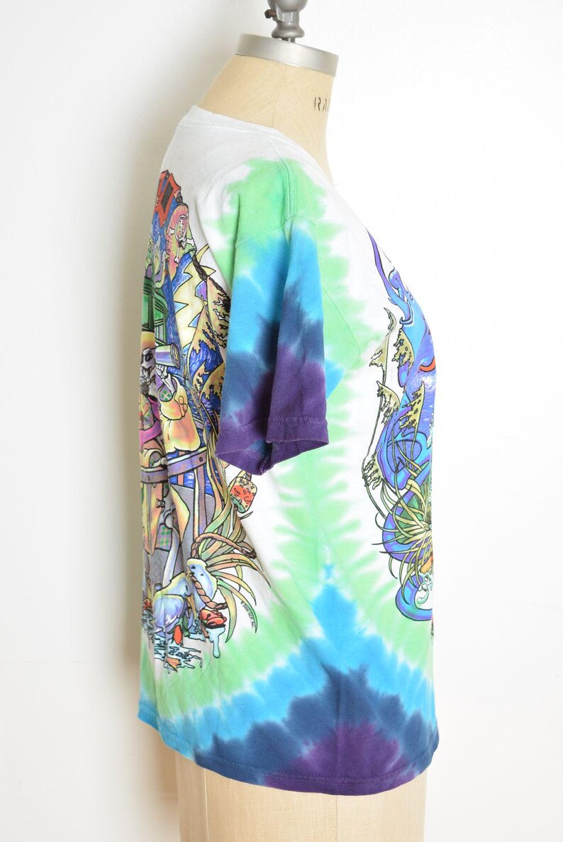 May include: A white, tie-dye t-shirt with a colorful graphic print. The shirt features a vibrant design with shades of blue, green, and purple. The graphic includes detailed illustrations. The shirt is displayed on a mannequin.