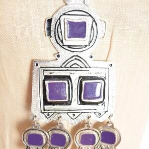 May include: A silver necklace with a large pendant featuring a geometric design with purple accents. The pendant has a square shape with two smaller squares inside, and the necklace has five smaller purple squares hanging from the bottom.